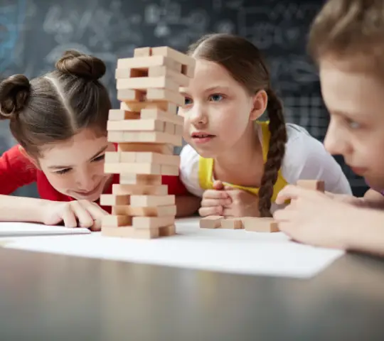 Children focus intently on a tall Jenga tower made of wooden blocks, surrounded by sheets of paper on a table.